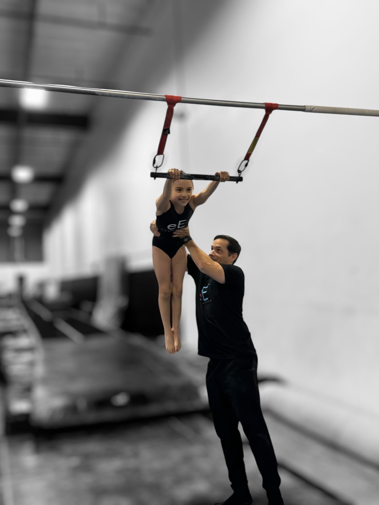 Gymnast performing on a high bar with a coach assisting in a gym setting.